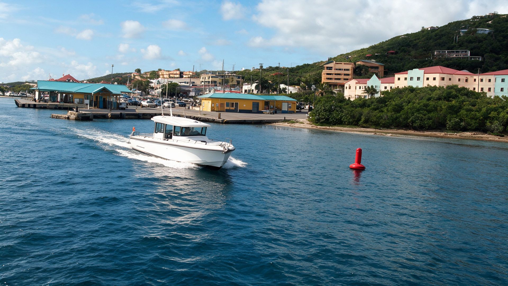 A white enclosed-cockpit water taxi arriving in Red Hook, St Thomas, past a red channel buoy, with the ferry dock, pastel buildings and green hillside ahead