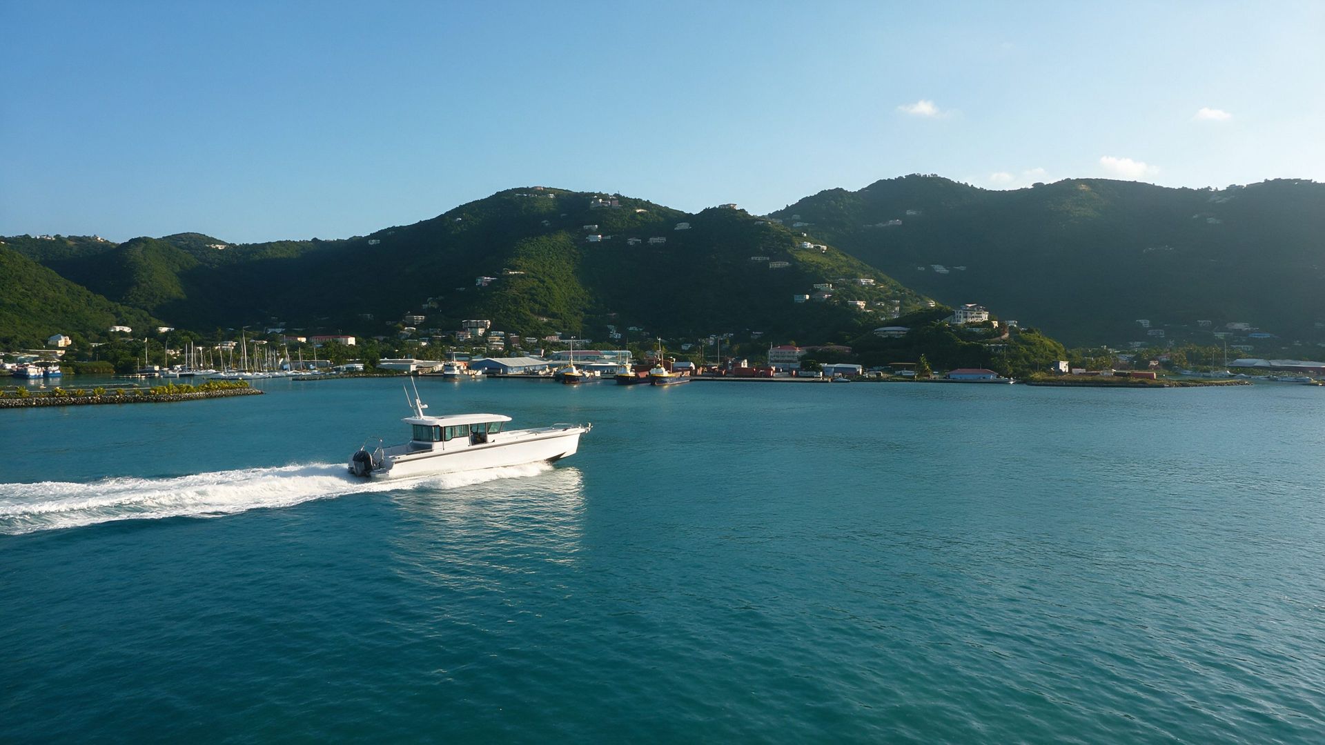 A white enclosed-cockpit water taxi crossing Road Harbour toward the Road Town waterfront and marinas, with the green hills of Tortola rising behind