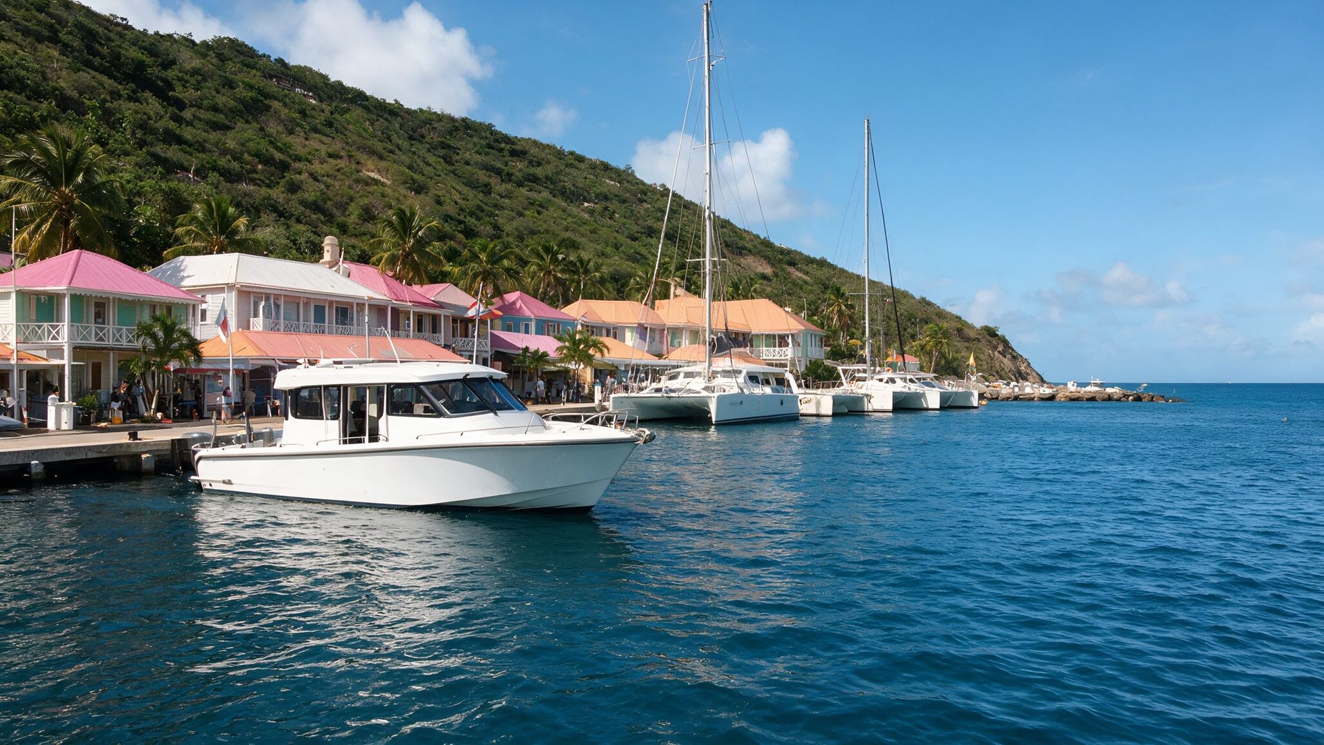 A white enclosed-cockpit water taxi alongside the wharf at Soper's Hole, West End, Tortola, in front of pastel pink, white and orange-roofed waterfront buildings and moored catamarans below a green hillside